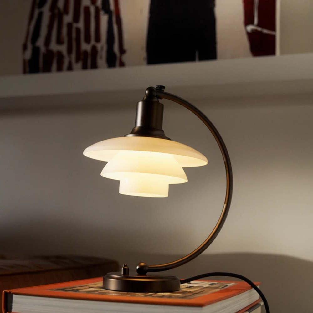 PH 2/2 Luna Aged Brass Opal Bordlampe i bruneret messing har Poul Henningsens karakteristiske treskærmssystem med skærme af mundblæst opalhvidt glas, der er blankt på ydersiden og sandblæst mat på undersiden. Det giver både et perfekt nedadrettet lys og et blødt, diffust lys rundt om lampen, som skaber stemning.