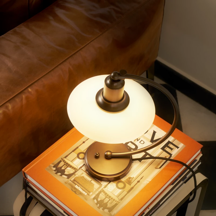 PH 2/2 Luna Aged Brass Opal Bordlampe i bruneret messing har Poul Henningsens karakteristiske treskærmssystem med skærme af mundblæst opalhvidt glas, der er blankt på ydersiden og sandblæst mat på undersiden. Det giver både et perfekt nedadrettet lys og et blødt, diffust lys rundt om lampen, som skaber stemning.